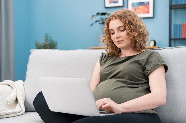 Beautiful pregnant woman using a laptop while working at cozy\
home girl typing something on laptop