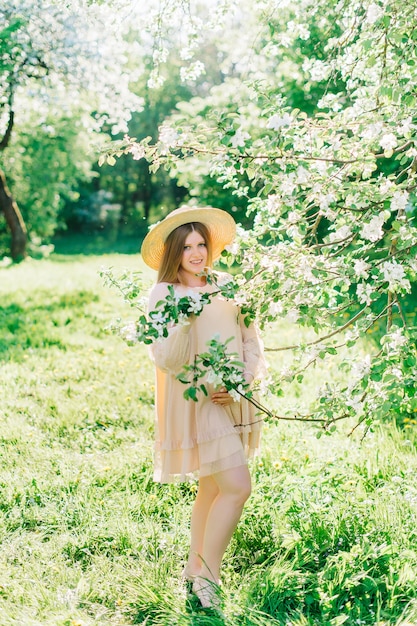 Beautiful pregnant woman wearing big hat and in blooming garden. Spring nature sunny day. Summer fashion concept.