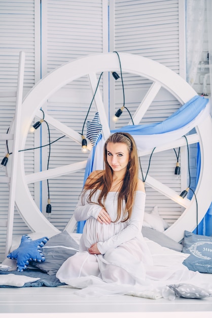 Beautiful pregnant woman in a white dress sits between the stylish cushions against the backdrop of large white wheel with light bulbs and wooden screens and holding pregnant belly