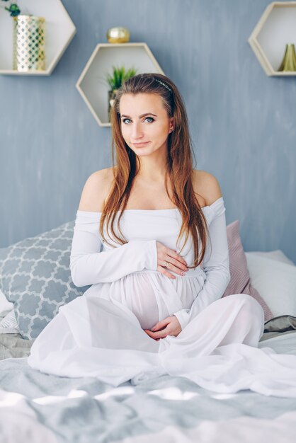 Beautiful pregnant woman in a white dress sitting on the white bed and holding on pregnant belly