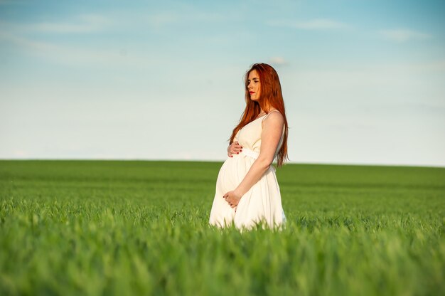 Красивая беременная женщина в белом платье гуляет в зеленом поле