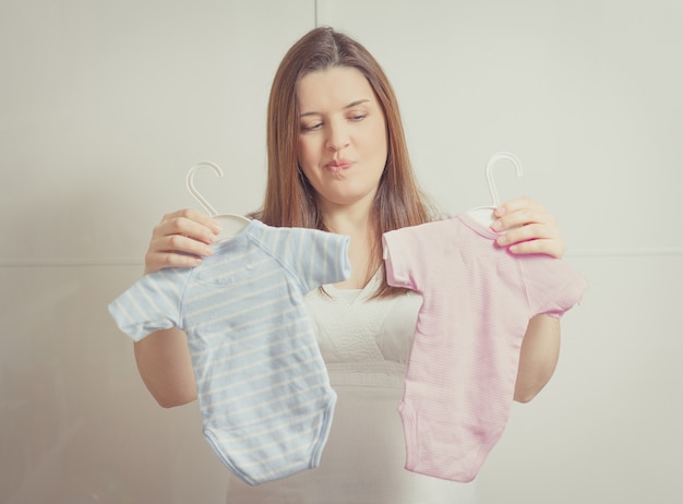 Beautiful pregnant young woman holding up bodysuits  for a girl or a boy newborn in soft pastel colors