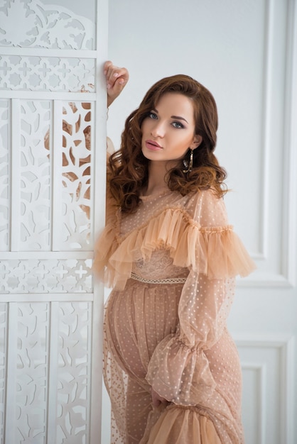 A beautiful pregnant young woman stands in a beige vintage dress near a white lace screen and touches the tummy with a baby.