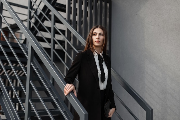 Beautiful professional business woman in a fancy black suit with a shirt tie and coat walks down the metal stairs outside in an office building