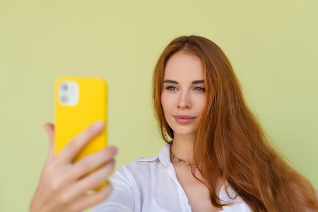 緑の背景にカジュアルなシャツを着た美しい赤い髪の女性は、スマートフォンで写真を撮って幸せです