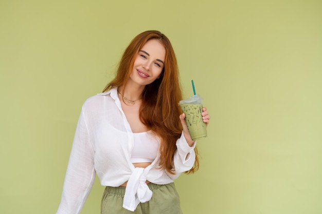 Красивая рыжеволосая женщина в повседневной рубашке на зеленом фоне позитивно наслаждается свежим ледяным зеленым чаем маття с молоком в жаркий летний день