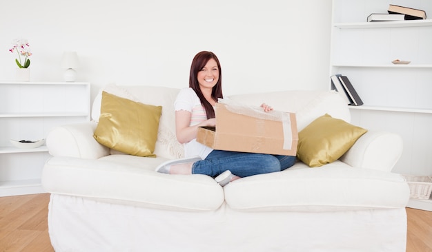 Beautiful red-haired female opening a carboard box while sitting on a sofa