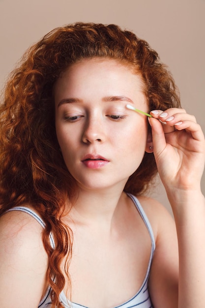 La bella ragazza dai capelli rossi pulisce la pelle dal trucco usando un batuffolo di cotone