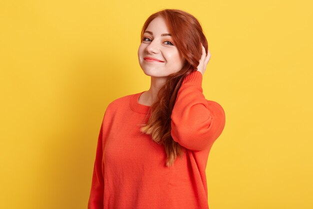 Beautiful red haired girl looking  with pleasant satisfied smile, keeping hand on her hair, wearing casual orange jumper, posing isolated over yellow wall.