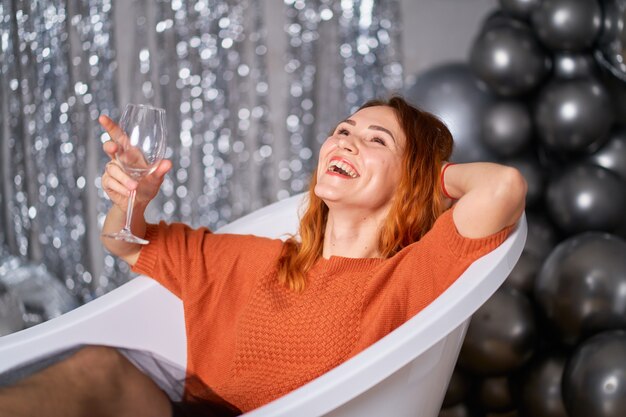 The beautiful red-haired girl rejoices sitting dressed in the bath. against the background balloons