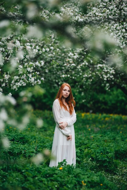 Beautiful red-haired girl in a white dress among blossoming apple-trees in the garden.