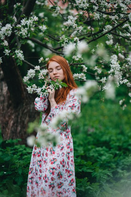 Beautiful red-haired girl in a white dress among blossoming apple trees in the garden 