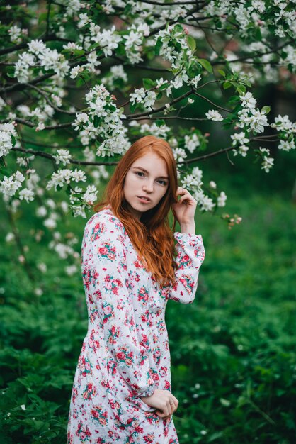 Beautiful red-haired girl in a white dress among blossoming apple trees in the garden 