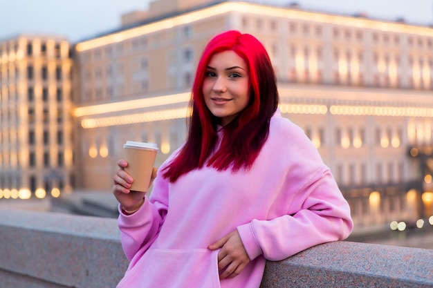 Красивая рыжеволосая девочка-подросток в розовой толстовке с капюшоном пьет кофе вечером на освещенной городской улице.
