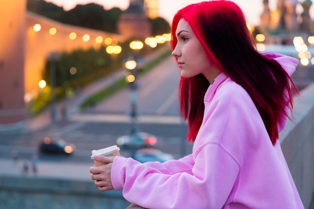 Bella ragazza adolescente dai capelli rossi in felpa con cappuccio rosa che beve caffè la sera sulla strada illuminata della città.