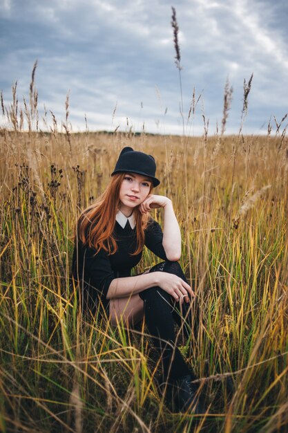 Beautiful red-haired woman in a black dress walks on an autumn field. 