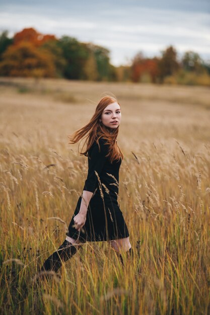 Beautiful red-haired woman in a black dress walks on an autumn field. 