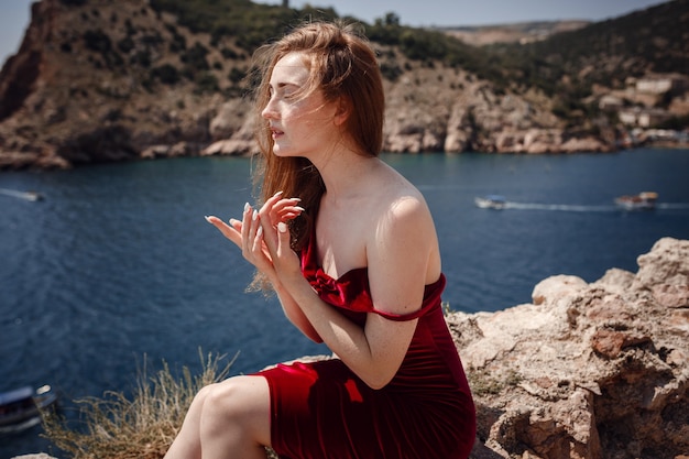 A beautiful red-haired woman in red dress is sitting on a rock with a gorgeous view of the seascape. Summer afternoon enjoyment of freedom and solitude