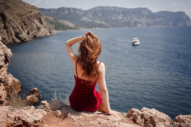 Красивая рыжеволосая женщина в красном платье сидит на скале с шикарным видом на морской пейзаж. Летний полдень наслаждение свободой и уединением