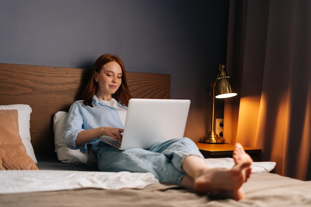 Beautiful red-haired young woman using laptop computer lying on\
bed at cozy dark bedroom. concept of leisure activity red-haired\
female at home during self-isolation.