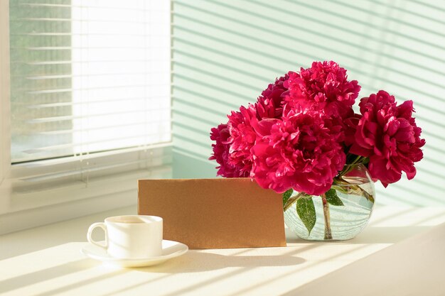 Beautiful red peonies tea coffee cup and letter on a light
background with hard shadows by the window selective focus