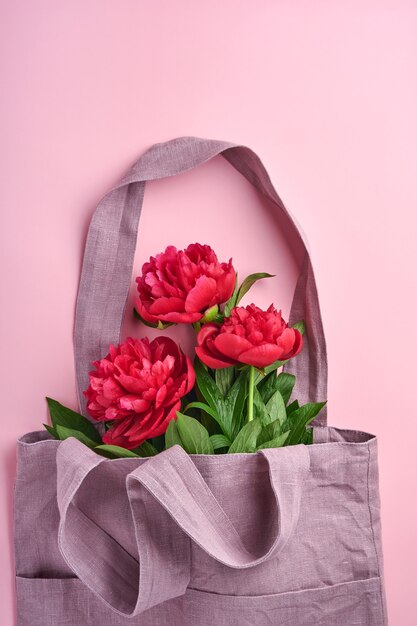 Beautiful red peony flowers bouquet in cotton eco tote bag over pink background, top view, copy space, flat-lay. Valentines day, mothers day background.