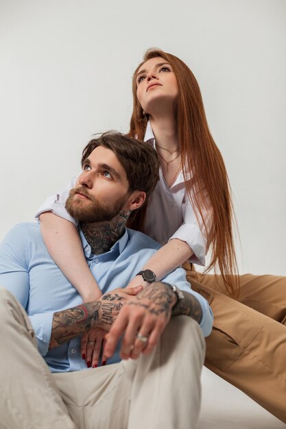 Beautiful redhaired girl and a handsome hipster man with a tattoo and a beard in a fashionable shirt are sitting in the studio Fashion stylish couple