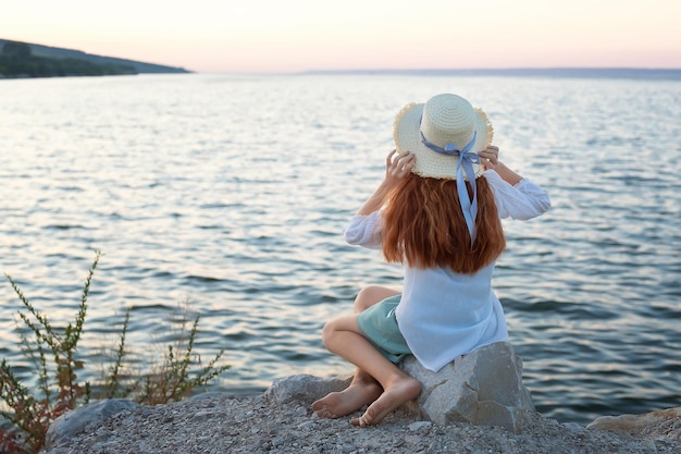 A beautiful redhaired girl in a hat sits on a stone on the banks of a river lake sea The girl is alone on the street The girl is sitting on the rocks