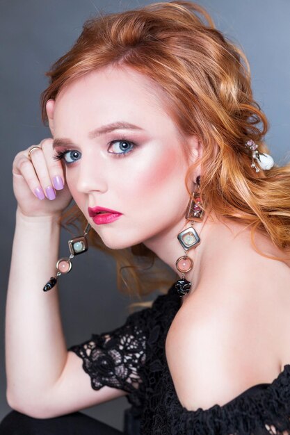 A beautiful redhaired girl with wavy hair makeup bare shoulders and long earrings posing on a gray background in the studio woman looking at camera