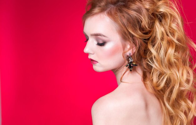 Beautiful redhaired girl with wavy hair and makeup posing on a red background in the studio in handmade earrings