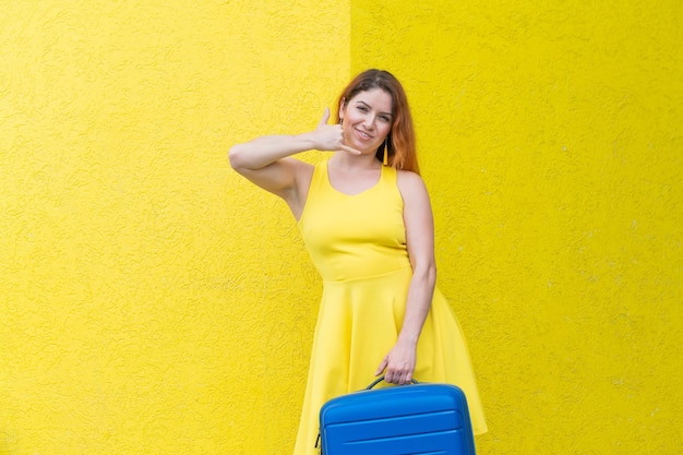 A beautiful redhaired woman in a dress holds a blue suitcase and gestures a phone call on a yellow background