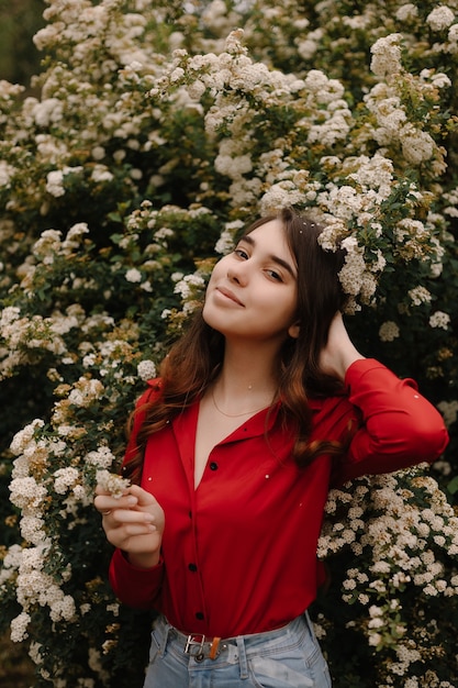 Красивая рыжеволосая девушка в красной блузке весной рядом с цветущими кустами