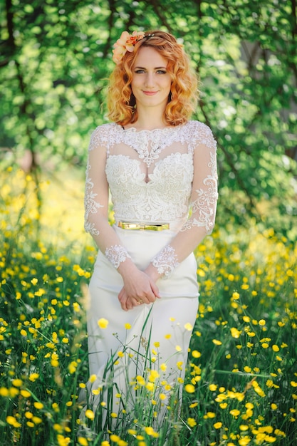 Красивая рыжая невеста в стильном свадебном платье в цветущем весеннем саду. Привлекательная рыжеволосая девушка в белом кружевном платье гуляет в зеленом парке.