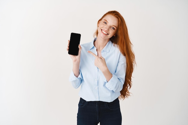 Beautiful redhead girl demonstrating mobile application pointing finger at blank smartphone screen standing over white background