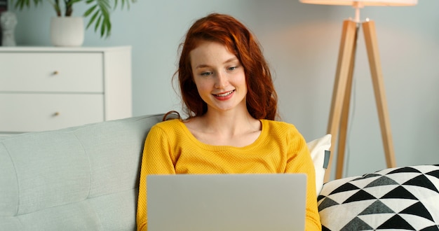 Beautiful redhead girl using silver laptop while sitting on sofa in living room at home