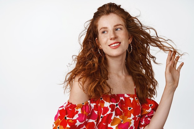 Beautiful redhead girl with makeup and stylish hairstyle playing with curly hair strand gazing aside and smiling dreamy standng in dress against white background