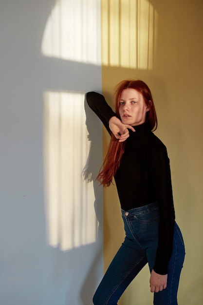 beautiful redhead long-haired girl dressed in black top and jeans is posing against the wall 