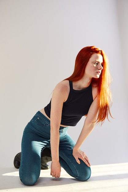 beautiful redhead long-haired girl dressed in black top and jeans is posing on the white background 