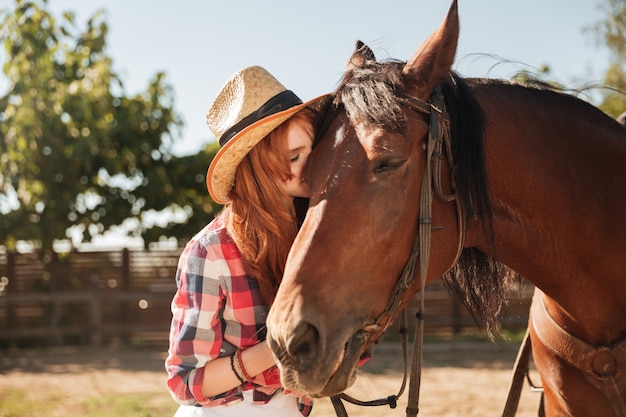 Beautiful redhead young woman cowgirl standing and kissing her horse