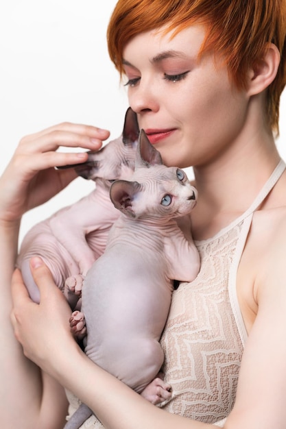 Beautiful redhead young woman gently hugging two purebred canadian sphynx cat to her chest and looki