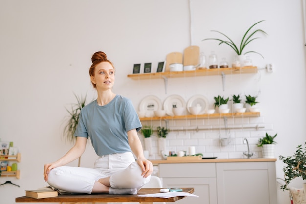 Beautiful redhead young woman sitting in lotus pose at the desk and looking away