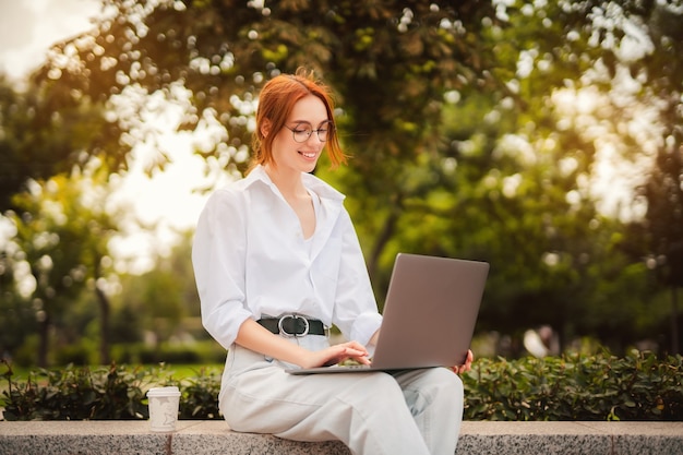 Beautiful redhead young woman sitting in the park and using laptop student university freelance wear...