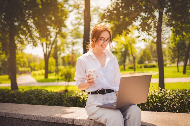 Beautiful redhead young woman sitting in the park and using laptop student university freelance wear...