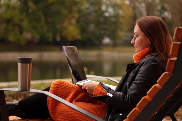 Photo beautiful redhead young woman sitting in the park and using laptop student university freelance wearing in glasses coffee to go looking in laptop