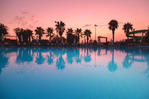 Beautiful reflection in swimming pool at colorful sunset Purple sky reflected in water palm trees sun beds umbrellas at night in summer Luxury resort Landscape with empty pool in twilight