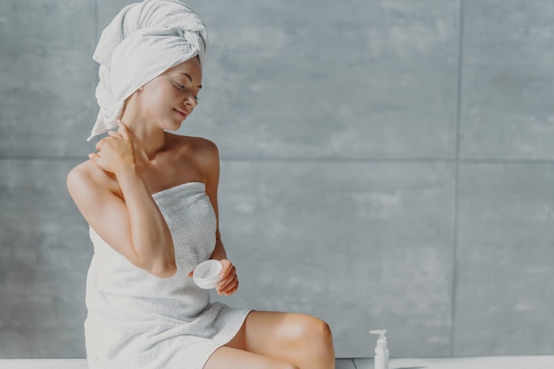 Красивая расслабленная женщина, обернутая полотенцем, применяя крем для лица