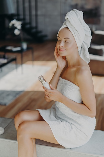 Beautiful relaxed woman wrapped in towel applying face cream