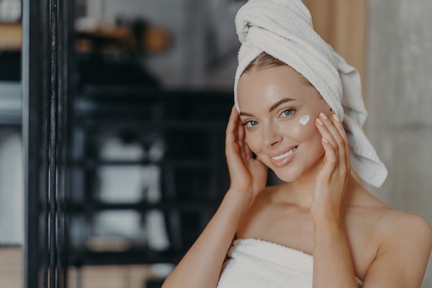 Beautiful relaxed woman wrapped in towel applying face cream