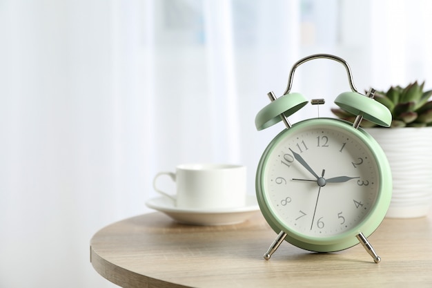 Beautiful retro alarm clock with cup of coffee and succulent plant on table against light