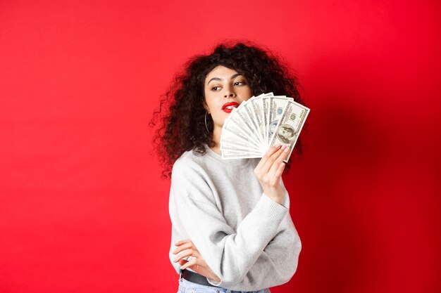 Beautiful rich woman looking sensual aside, waving at herself with dollar bills fan, standing seductive on red wall.